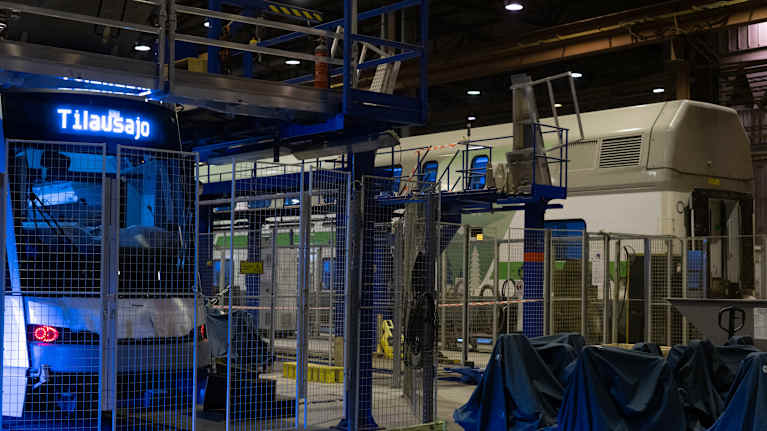 Raitiovaunu verkkoaidan takana tehdashallissa. Keulassa ylhäällä näytössä lukee Tilausajo. Taustalla näkyy kaksikerroksinen VR:n junavaunu.