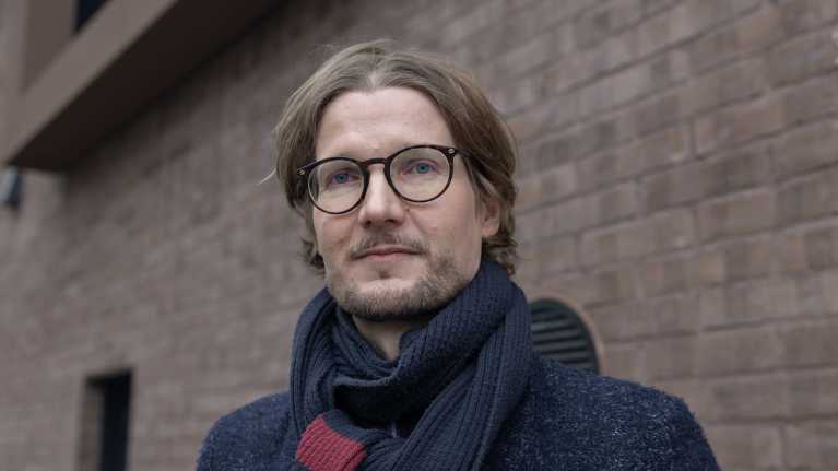 Silmälasipäinen mies kaulahuivi kaulassaan seisoo tiilisen seinän edustalla.