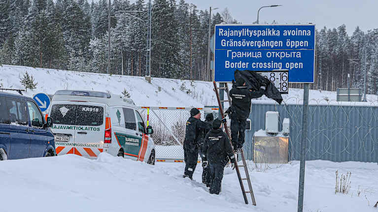 Kolme rajamiestä seisoo lumihangessa ja peittää mustalla muovisäkillä kylttiä, jossa lukee Vartiuksen rajanylityspaikan aukioloajat.