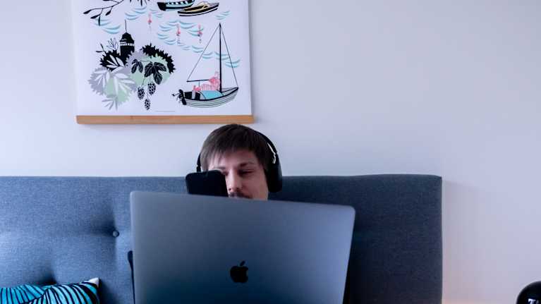 Henkilö istuu sängyllä luurit päässä katsoen puhelintaan ja kannettava tietokone sylissä.