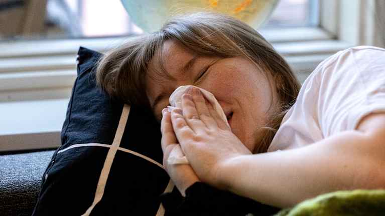 Nainen makaa sohvalla viltin alla ja niistää nenäänsä.