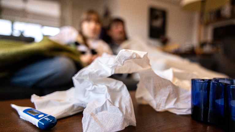 Nainen ja mies makaa sohvalla vilttien alla, etualla pöydällä käytettyjä nenäliinoja ja kuumemittari.