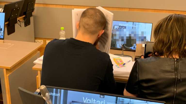 Miehen takaraivo. Mies peittelee kasvojaan paperilla.