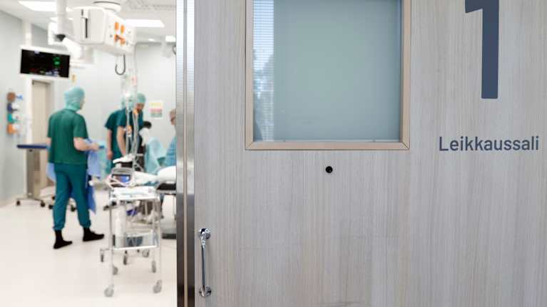 Raollaan oleva liukuovi, jossa on pieni ikkuna ja teksti leikkaussali. Raosta näkyy leikkaussalivaatteisiin pukeutuneita ihmisiä ja salin välineitä.