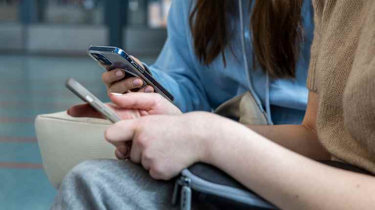 Kaksi henkilöä istuu vierekkäin ja katselee älypuhelimiaan. He vaikuttavat olevan rentoja ja keskittyneitä puhelimiensa käyttöön.