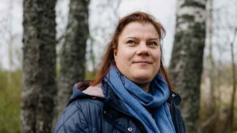 Nainen seisoo metsässä kevättakin ja huivin kanssa. Taustalla näkyy koivunrunkoja.