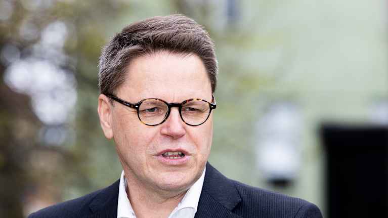 Henkilö kuvassa seisoo ulkona, yllään tumma puku ja vaalea kauluspaita.