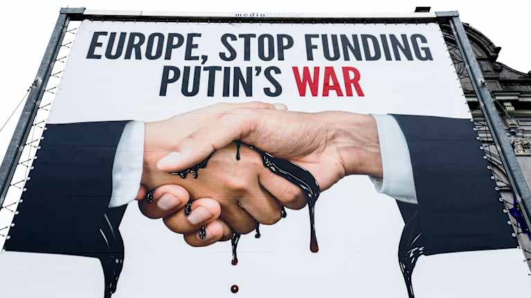 Suuri banderolli, jossa kätellään, käsien välistä tihkuu mustaa öljyä. Yläpuolella lukee Europe, stop funding Putin's war.