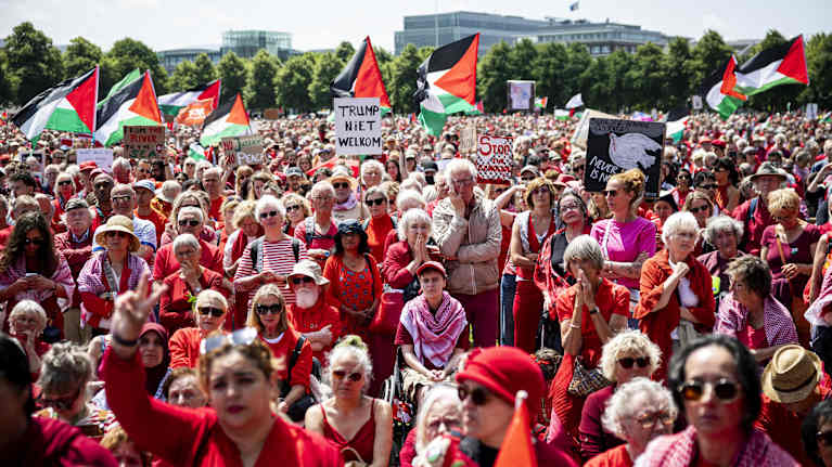 Ryhmä ihmisiä pukeutuneina punaiseen osallistuu mielenosoitukseen, jossa on esillä Palestiinan lippuja ja kylttejä, kuten "Trump niet welkom" ja "Stop the genocide."
