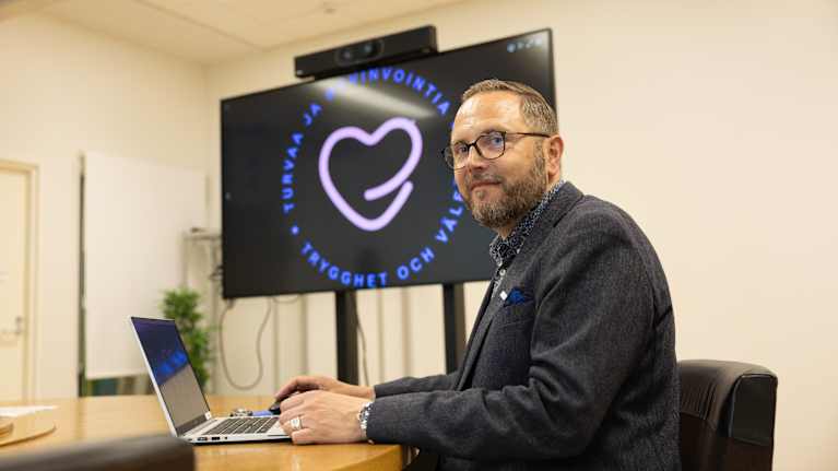 Mies istuu pöydän äärellä kannettavan tietokoneen kanssa ja katsoo kameraan.