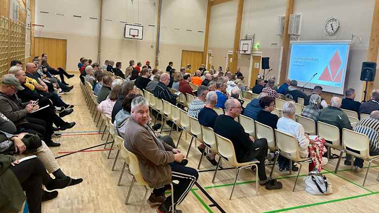 Tilaisuus käynnissä liikuntasalissa, jossa useat osallistujat seuraavat esitystä projektorikankaalla.