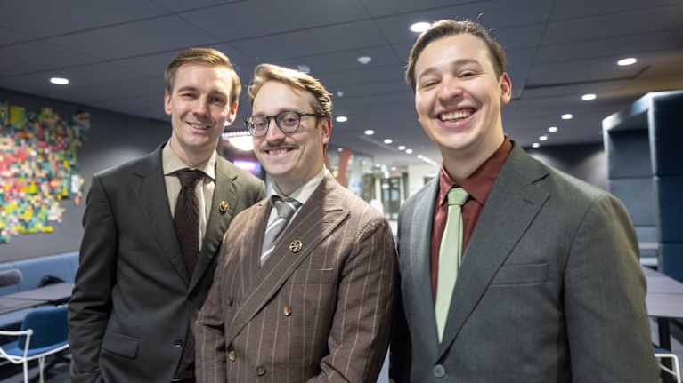 Kolme tyylikkäästi pukeutunutta henkilöä poseeraa sisätiloissa. Taustalla näkyy värikäs seinätaideteos ja modernia sisustusta.