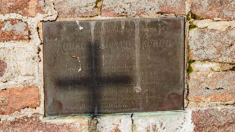Kuvassa on metallinen muistolaatta, joka on kiinnitetty punatiiliseen seinään.