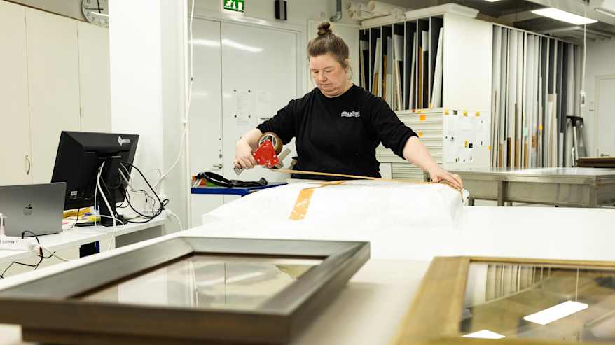 Museotyöntekijä pakkaa taideteosta kuljetusta varten, käyttäen kuplamuovia ja teippiä. Etualalla on toinen teos odottaa käsittelyä.