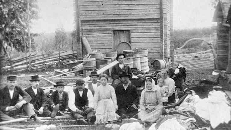 Vanha ryhmäkuva, jossa istuu useita ihmisiä maalaismaisemassa torppa taustallaan.