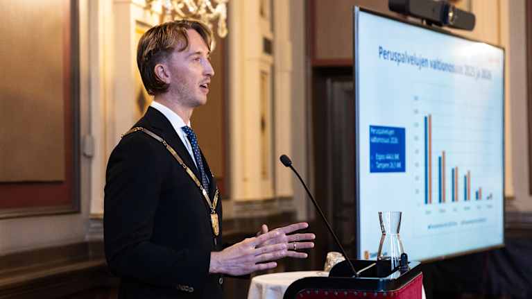 Henkilö seisoo puhujakorokkeella muodollisessa ympäristössä, pukeutuneena tummaan pukuun ja kantaa kaulassaan arvoketjua.