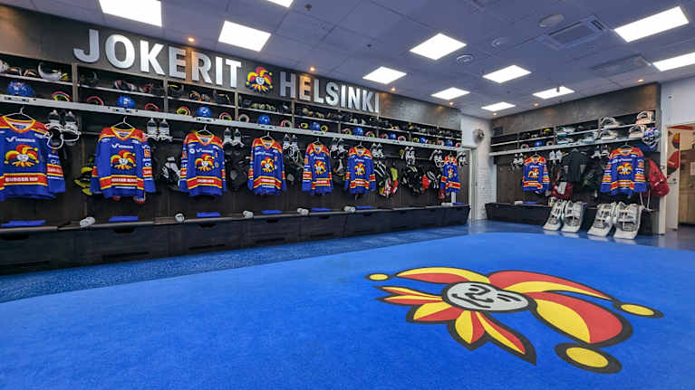 Jokerien pukuhuone, jossa sininen lattia ja jokerien logo lattiassa. Seinällä hyllyissä jokerien pelipaitoja.