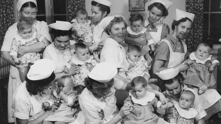 Mustavalkoinen kuva, jossa on useita hoitajia vauvoja sylissään.