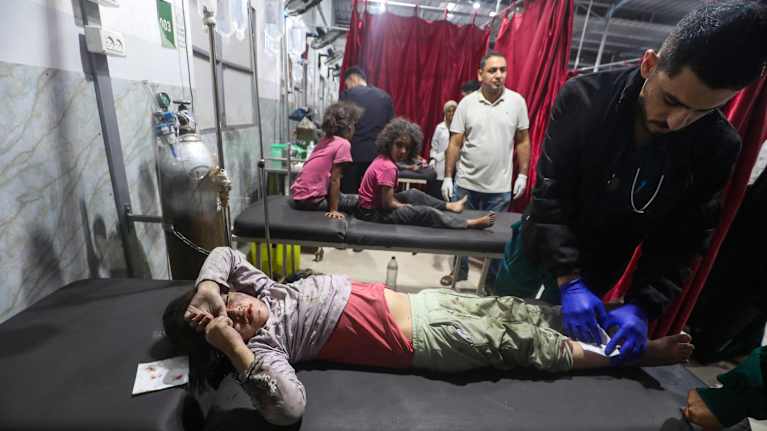 Hoitaja puhdistaa sairaspedillä makaavan lapsen haavoja. Taustalla enemmän sairaspetejä, joilla istuu lapsia.