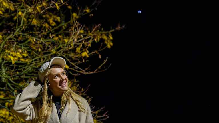 Nainen seisoo yöllä ulkona, puettuna vaaleaan takkiin, keltalehtisen puun edessä.