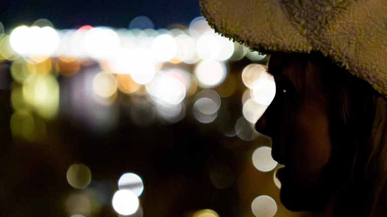 Pörrölippalakkisen naisen katse kohti kaupunkivalojen loistetta yön hämärässä.