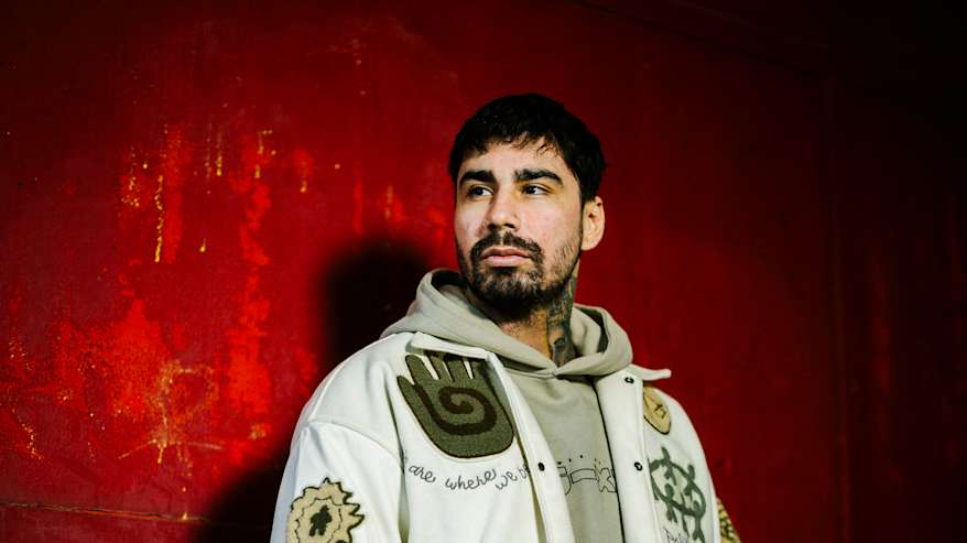 Henkilö seisoo syvää punaista taustaa vasten, pukeutuneena vaaleaan takkiin.