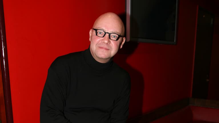 Lasse Norres istuu punaisella sohvan reunalla tummissa vaatteissa. Taustalla näkyy punainen seinä ja televisioruutu.