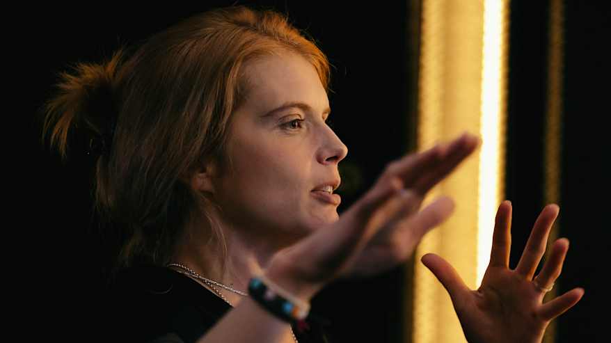 Alina kuvattuna kesken haastattelun sivusta, ilmetii käsillään. Taustalla lämmin valonkajo.