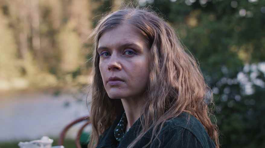 Pitkähiuksinen vaalea nainen katsoo kameraan vakavalla ilmeellä, taustalla järvimaisema.