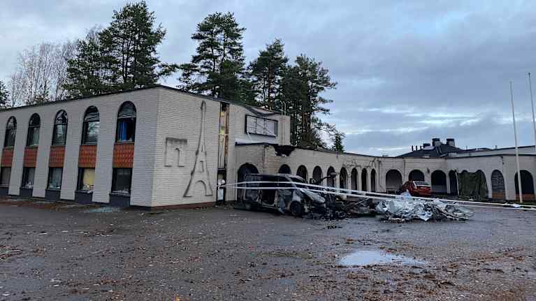 Vaalea rakennus, joka on vaurioitunut räjähdyksessä. Rakennuksen edustalla on pahoin vaurioitunut ja mustunut pakettiauto.