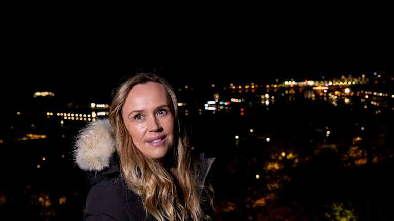 Nainen seisoo ulkona yöllä ja on pukeutunut lämpimään talvitakkiin. Taustalla näkyy kaupungin valot 