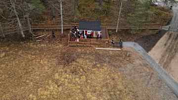 Ilmakuva vanhasta punaisesta saunasta ja sen terassilla olevista ihmisistä. Rakennus sijaitsee metsän reunassa rinteessä.