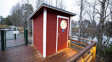 Punainen inva-wc, jossa on pyöreä ikkuna, sijaitsee puisella laiturilla veden äärellä.