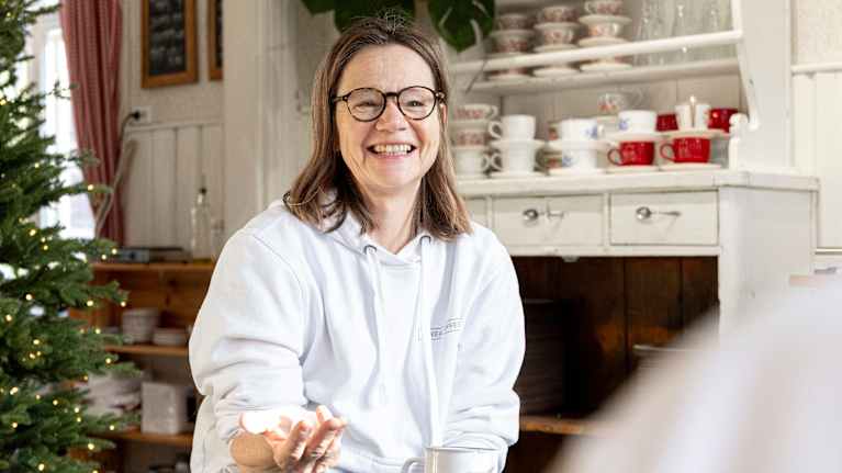 Kuvassa henkilö istuu kahvilassa pitäen mukia kädessään, taustalla hyllyillä on astioita ja joulukuusi.