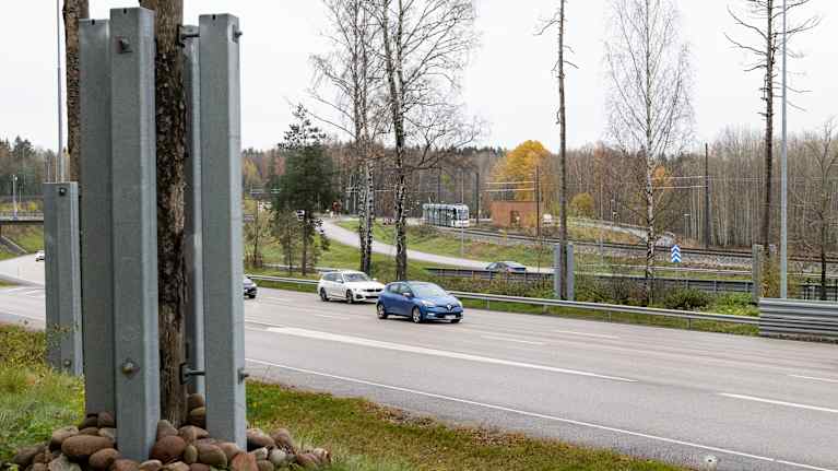 Metallitolpilla tuettuja osittain kelottuneita männyn runkoja tien reunassa ja eri suuntaa menevien teiden välissä. Taustalla raitiovaunu.