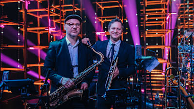 Kaksi muusikkoa poseeraamassa saksofonin ja trumpetinin kanssa värikkäässä lavaympäristössä.