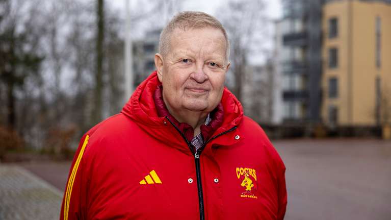 Mies ulkona punaisessa takissa. Taustalla näkyy syksyistä kerrostaloaluetta. 