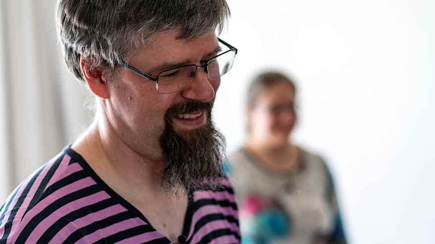 Paiste Mäenpää seisoo huoneessa, ja taustalla näkyy toinen henkilö. Etualalla olevan henkilön vaatteet ovat värikkäitä ja raidallisia. 