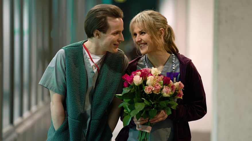 Mies ja nainen sairaalamiljöössä. Naisella ruusukimppu kädessä