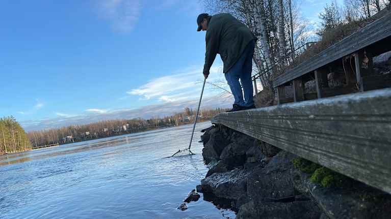 Mies lippoaa haavilla laiturilla ja kalastaa jokimaisemassa.
