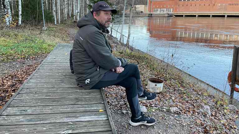 Mies istuu puisella laiturilla joen rannalla syksyisessä maisemassa. Taustalla näkyy suuri tiilirakennus ja metsää.