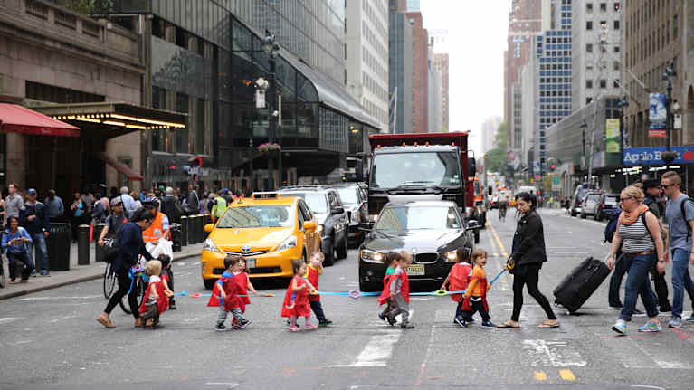 Aikuiset johdattavat pieniä lapsia monikaistaisen autotien yli suojatietä pitkin. Lapsilla on punaiset liivit ja he pitävät kiinni sinisestä nauhasta.