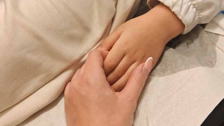 Äiti ja lapsi pitävät toisiaan kädestä kiinni. Kuvassa aikuisen ja lapsen käsi sairaalapedillä.