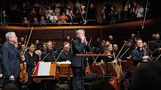 Kapellimestari kiittää yleisöä konsertin jälkeen. Yleisö seisoo taustalla, orkesteri seisoo.