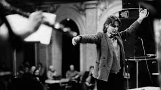 Nuori kapellimestari johtaa orkesteria korokkeella. Yleisö seuraa esitystä taustalla.