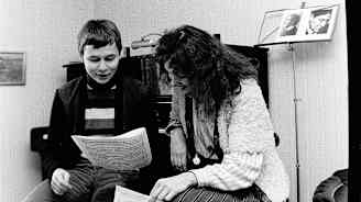 Nuori mies ja nainen katselevat hymyillen nuottipaperia pianon ja nuottitelineen edessä.