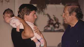 Kuvassa kaksi aikuista keskustelevat keskenään. Toinen aikuisista pitelee olkapäällään pientä vauvaa, joka vaikuttaa uteliaalta ympäristöstä.