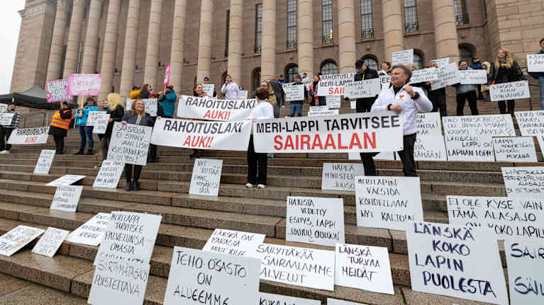 Paljon kylttejä eduskuntatalon portailla. Taustalla mielenosoittajia.