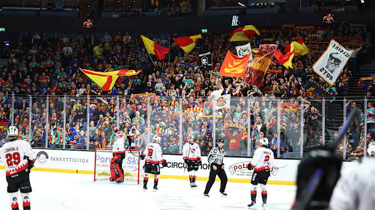 Jääkiekko-ottelu käynnissä ja innokkaat kannattajat heiluttavat lippuja katsomossa kannustaen joukkuettaan.