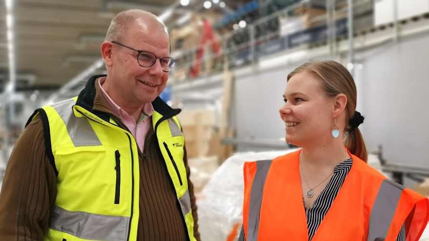 Turvaliiveihin pukeutuneet mies ja nainen keskusteevat hymyillen, taustalla tehtaan tuotantotiloja.